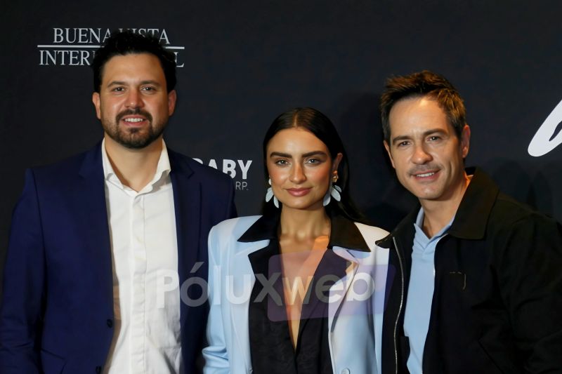 Fotografía de tres personas posando sonrientes de medio cuerpo frente a un fondo negro con letras blancas parciales. De izquierda a derecha aparece un hombre con barba vistiendo traje azul y camisa blanca desabotonada, una mujer al centro con blazer claro y aretes grandes, y un hombre a la derecha con chaqueta oscura y playera polo azul claro.