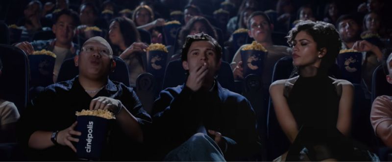 Tom Holland, Zendaya y Jacob Batalon en el spot de Cinépolis para "Spider-Man: Un Nuevo Día" Fotografía fija del spot de Cinépolis "Hechos de Cine". En una sala de cine oscura y con la iluminación ambiental de la pantalla, aparecen sentados de izquierda a derecha Jacob Batalon, Tom Holland y Zendaya disfrutando de una función. Jacob Batalon lleva lentes y sostiene un bote de palomitas con el logotipo de Cinépolis, al igual que Tom Holland, quien come palomitas con una mano, mientras que Zendaya observa hacia un costado con expresión seria y elegante vestimenta oscura. De fondo, otros espectadores disfrutan de la función.