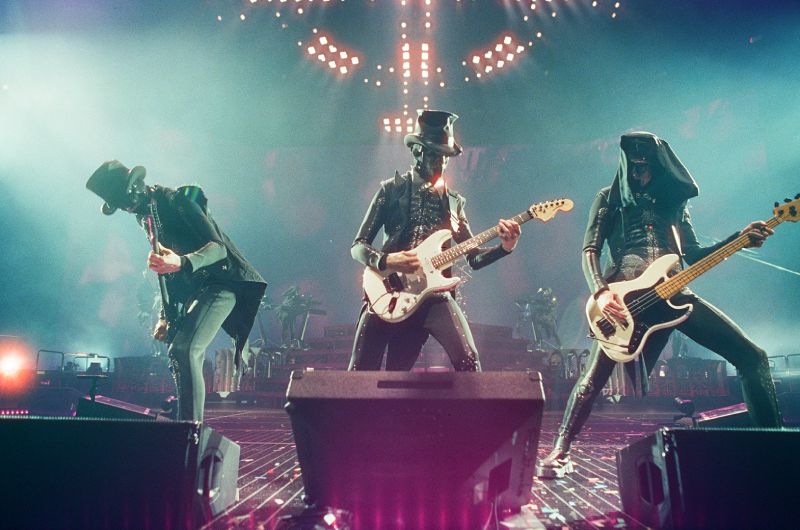 Fotografía en vivo del grupo de metal Ghost durante su presentación en concierto sobre el escenario del Palacio de los Deportes. Tres músicos pertenecientes a los Nameless Ghouls aparecen alineados tocando sus instrumentos bajo una iluminación escénica azul y verde con luces de fondo. Los músicos visten trajes oscuros de época con chaquetas estilizadas, sombreros de copa y máscaras metálicas detalladas. A la izquierda, un guitarrista inclina su instrumento; al centro, otro guitarrista sostiene una guitarra eléctrica blanca modelo Fender Stratocaster; a la derecha, un bajista con postura abierta toca un bajo eléctrico blanco Fender Precision Bass. En el suelo del escenario se observan monitores de retorno de audio negros y confeti esparcido.