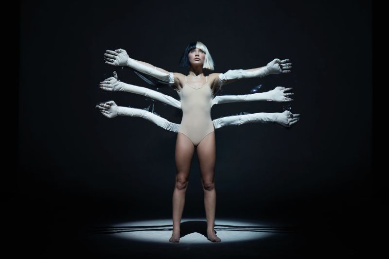 Sia Nostalgic for the Present Figura en escena iluminada por un reflector, acompañada por múltiples brazos artificiales que forman una composición simétrica.