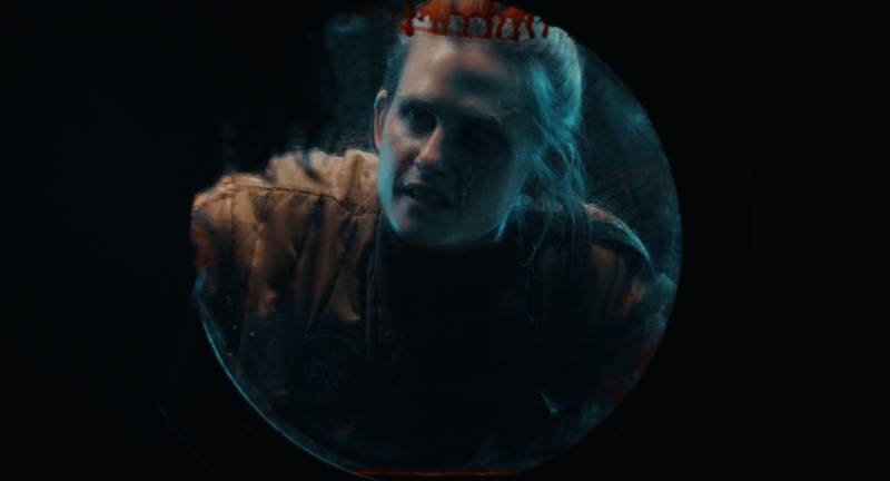 Fotografía de un personaje visto a través de una escotilla circular iluminada por un tono azulado, que refuerza la sensación de encierro y tensión en un entorno oscuro.