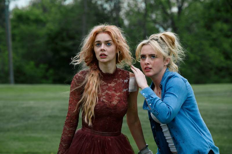 Dos mujeres observan con atención algo fuera de cuadro en un entorno natural, una de ellas con vestido rojo y la otra con camisa azul, ambas mostrando preocupación en su expresión.