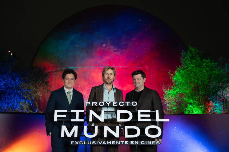 Presentación de Proyecto Fin del Mundo en Ciudad de México Tres integrantes del equipo de la película posan frente a una estructura iluminada con colores vibrantes, acompañados por un letrero promocional del filme en un entorno nocturno.