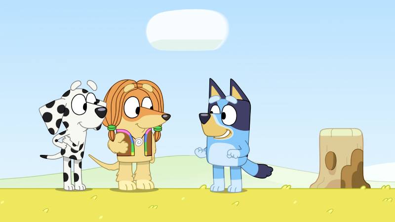 Tres personajes caninos animados conversan en un campo verde bajo un cielo despejado, transmitiendo un ambiente amistoso y alegre.