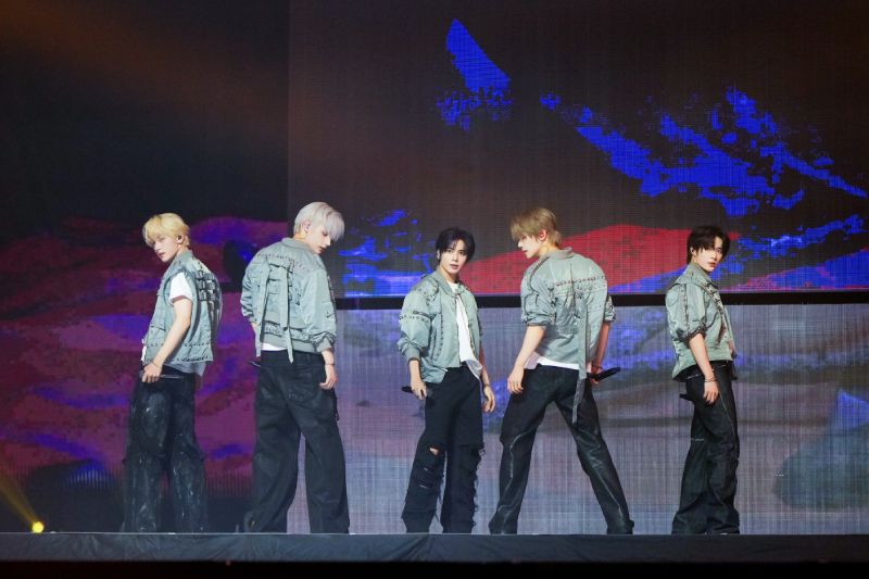 Los cinco integrantes del grupo de K-pop TOMORROW X TOGETHER posan de pie sobre un escenario de concierto vestidos con chaquetas grises estilo industrial frente a pantallas gigantes de fondo.   