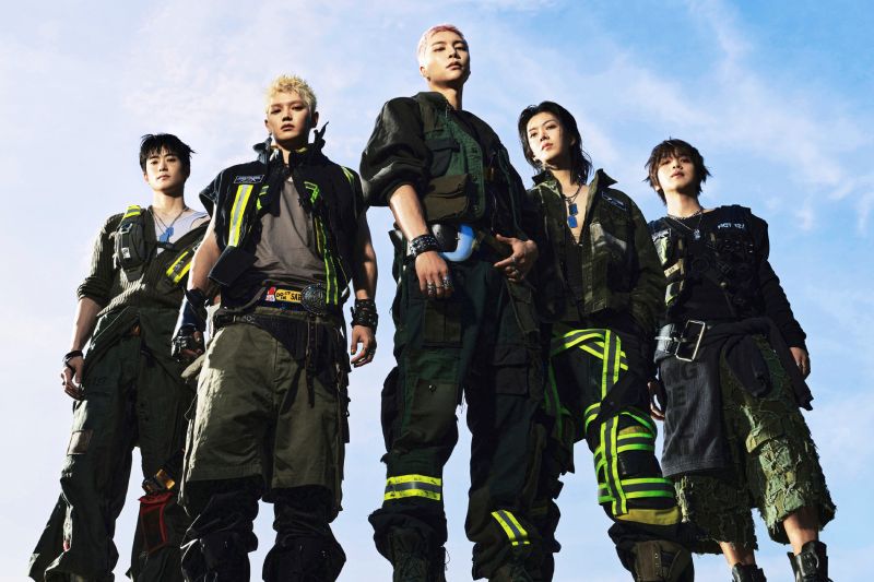 Promocional oficial del concierto "NCT 127 5TH TOUR ‘NEO CITY: SEOUL - THE REDLINE’ in CINEMAS". Fotografía promocional de los integrantes de la agrupación de K-pop NCT 127. Aparecen cinco miembros posando en contrapicado bajo un cielo azul despejado, luciendo atuendos de estilo industrial y táctico en tonos verde oliva, negro y beige con cintas fluorescentes.