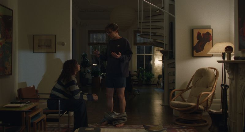 Dos personas en una sala cálida: una sentada con suéter a rayas y otra de pie frente a ella, con pantalones a la altura de los tobillos. El entorno muestra muebles de madera, una escalera de caracol y decoración hogareña.