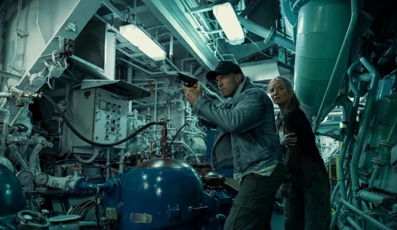 Jason Statham en la sala de máquinas en Código: Venganza Jason Statham apuntando con una pistola dentro de la sala de máquinas de un barco mientras protege a una mujer detrás de él.