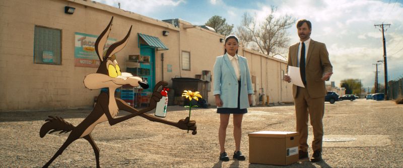 El Coyote sosteniendo una flor y una lata de spray de broma en un callejón frente a Lana Condor y Will Forte.