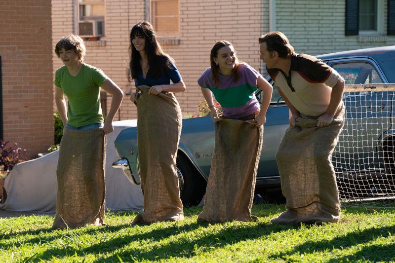 Escena retrospectiva o cotidiana que muestra a Anne Hathaway, Ewan McGregor y sus dos hijos riendo y compitiendo alegremente en una carrera de costales en el jardín de su casa.