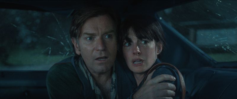 Plano medio cerrado desde el interior de un automóvil con los cristales estrellados donde Ewan McGregor abraza con tensión a Anne Hathaway mientras ambos observan aterrados hacia el exterior.