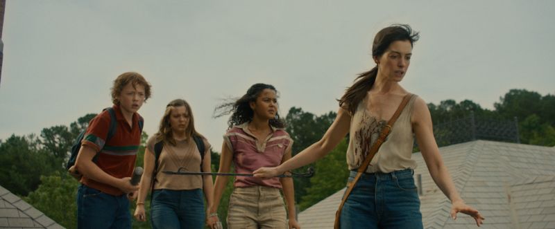 Fotograma de Anne Hathaway al frente con una vara de metal ensangrentada guiando a tres jóvenes sobre el tejado de una vivienda mientras observan alerta su entorno.