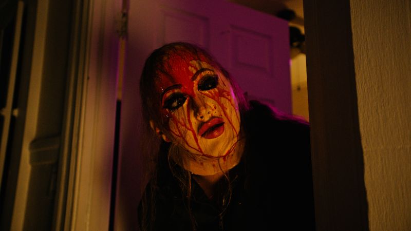 Frankie acechando en las sombras Fotograma en plano medio en penumbra con luz morada y cálida, donde un personaje asoma la cabeza por el marco de una puerta vistiendo una máscara femenina manchada con abundante sangre roja en la frente.