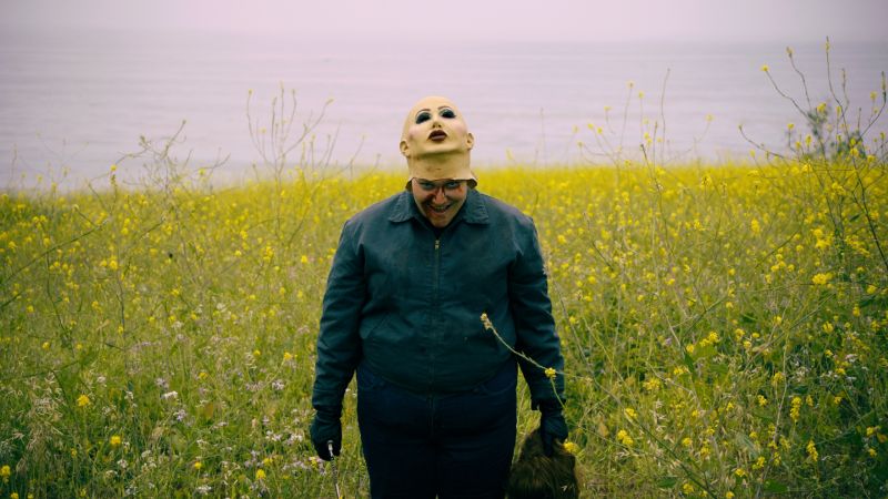 Frankie en el campo de flores Plano general de un personaje en medio de un campo de flores amarillas bajo un cielo nublado, vistiendo una chamarra oscura y una máscara de manicura pálida levantada hacia la frente, revelando una sonrisa desquiciada y sangrienta.