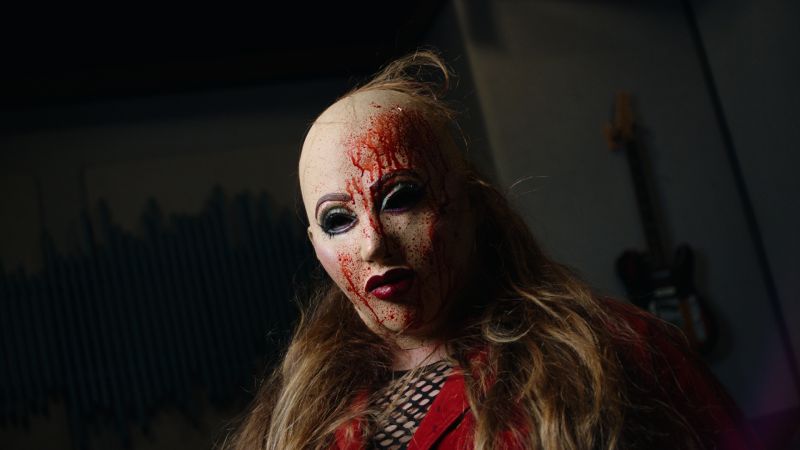 Frustración musical y locura en Frankie Maniac Woman Primer plano en un estudio de grabación con guitarras al fondo, mostrando a la protagonista con peluca rubia y una máscara con salpicaduras de sangre cayéndole por el rostro.