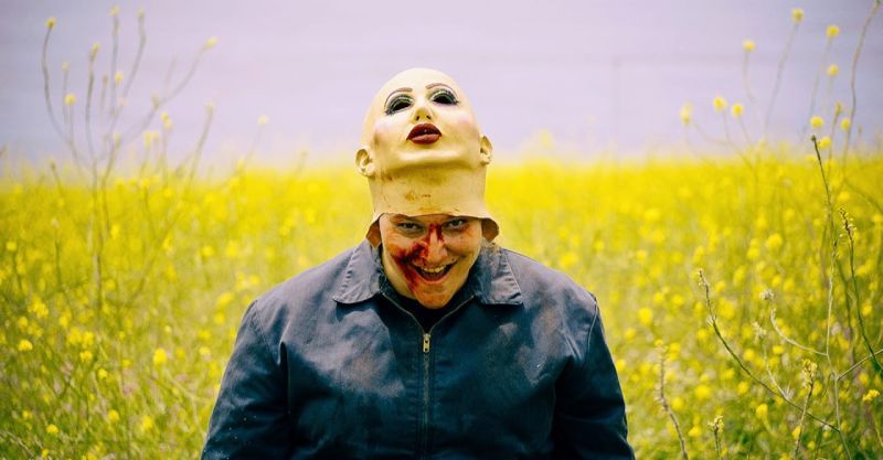 La revelación del monstruo en Frankie Maniac Woman Plano medio corto en un campo abierto donde la persona sonríe con la boca manchada de sangre mientras mantiene la máscara de látex levantada sobre la cabeza a la altura de la frente.