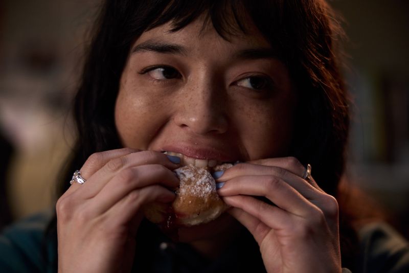 Primer plano de Hanna (Midori Francis) mordiendo una dona de mermelada cubierta de azúcar glass con una expresión de ansiedad y placer contenido.