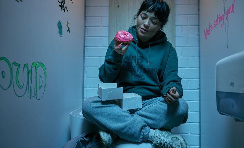 Hanna (Midori Francis) sentada con las piernas cruzadas sobre un inodoro en el cubículo de un baño, sosteniendo una dona rosada con chispas extraída de una caja de repostería.