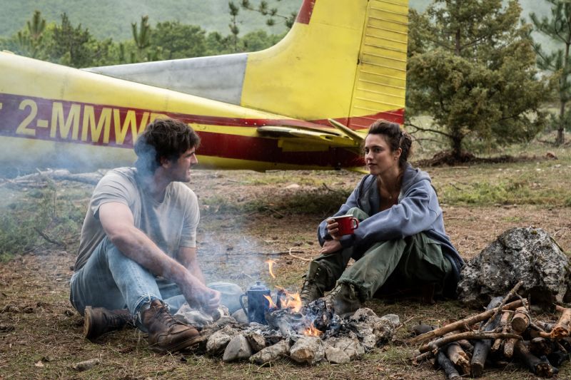 Hig y Cima junto a la fogata en La Guerra de los Últimos Jacob Elordi como Hig y Margaret Qualley como Cima sentados junto a una fogata al aire libre platicando con la avioneta amarilla de fondo.