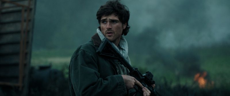 Jacob Elordi como Hig en combate en La Guerra de los Últimos Jacob Elordi en el rol de Hig mirando atentamente a su alrededor mientras sostiene un rifle de asalto en un entorno sombrío.