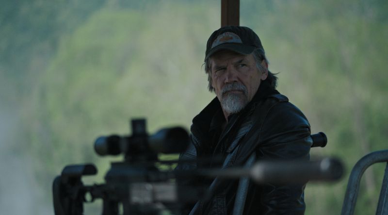 Josh Brolin como Bangley en posición de defensa Primer plano de Josh Brolin interpretando a Bangley con gorra y barba vigilando desde un puesto de observación tras un rifle con mira telescópica.