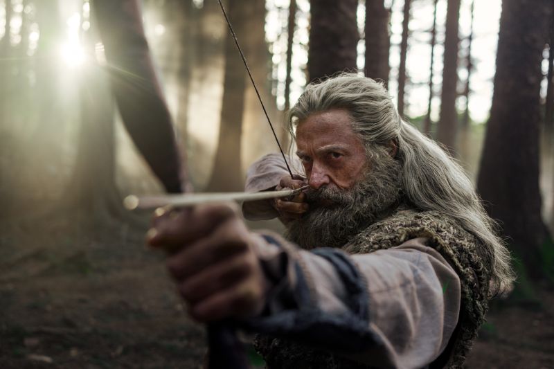 Hugh Jackman como Robin Hood apuntando su arco Primer plano de Hugh Jackman caracterizado como Robin Hood apuntando cuidadosamente con un arco de madera y una flecha en medio del bosque.
