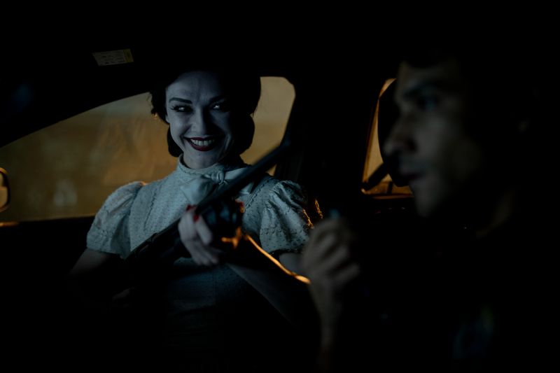 Espíritu aterrador en La Noche del Demonio: Están Entre Nosotros Un espíritu femenino con maquillaje pálido y sonrisa pertubadora sentada en el asiento trasero de un automóvil apuntando con un arma hacia el conductor.