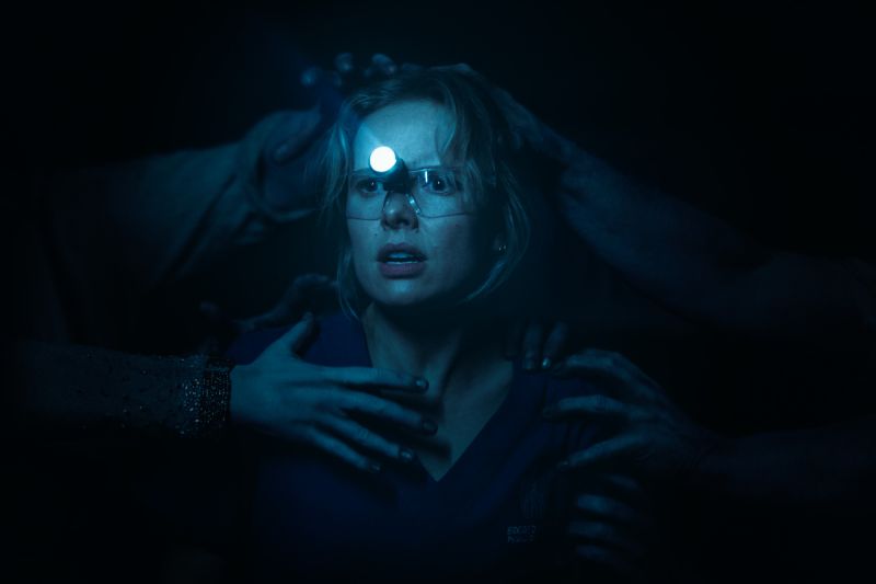 Gemma atrapada por entes espirituales Gemma con una lámpara frontal de dentista en la cabeza mientras múltiples manos espectrales y oscuras emergen de las sombras para sujetarla.
