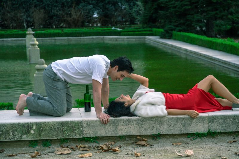 Aislinn Derbez y Mauricio Ochmann en escena de Hasta el fin del mundo Fotograma de Hasta el fin del mundo donde Aislinn Derbez yace acostada con un vestido rojo sobre el borde de una fuente de piedra mientras Mauricio Ochmann se inclina sonriente hacia ella.