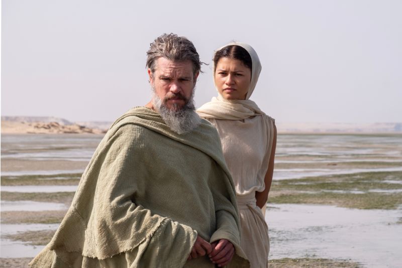 Fotograma de la película La Odisea donde se observa a Matt Damon con vestimenta clásica de la Grecia antigua y barba canosa representando a Odiseo, acompañado por Zendaya con túnica y velo blanco en una locación natural costera.