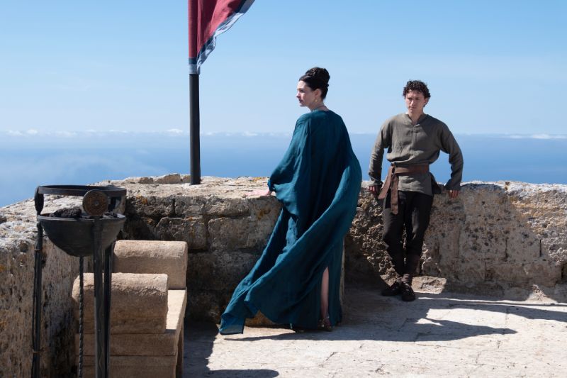 Escena del filme La Odisea con Anne Hathaway luciendo un vestido azul estilo griego y Tom Holland como su hijo Telémaco en la terraza de una fortaleza de piedra junto al mar.