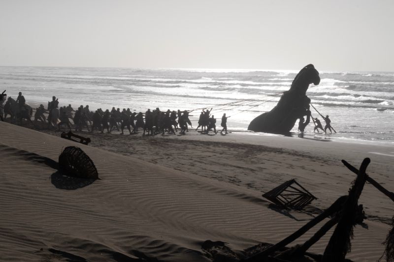 Captura cinematográfica en blanco y negro que muestra a decenas de hombres arrastrando la estructura del Caballo de Troya a lo largo de una playa durante una de las secuencias de flashback de la película.