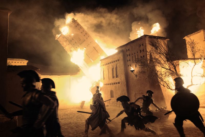 Plano de acción de La Odisea que capta a varios soldados con cascos griegos combatiendo en medio de un incendio nocturno con una torre cayendo en llamas tras ellos.