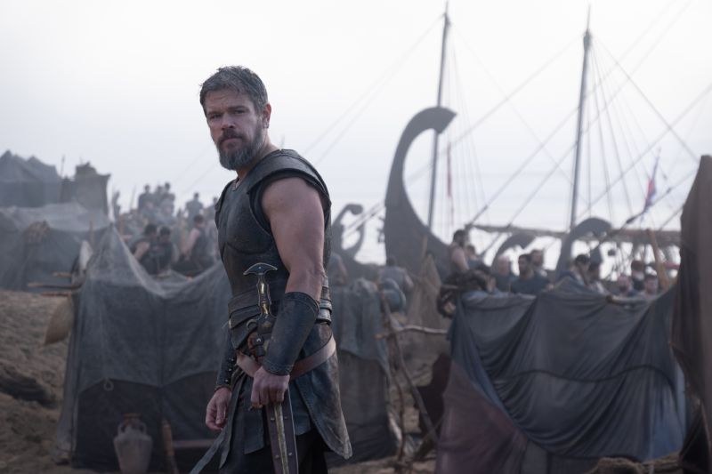 Fotograma de la película La Odisea donde aparece Matt Damon en primer plano portando armadura de combate antigua y sosteniendo una espada, situado en una playa repleta de tiendas de campaña y trirremes griegos.