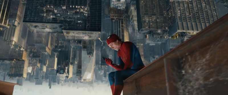Momento introspectivo de Peter Parker en las alturas de Nueva York Perspectiva invertida de Spider-Man con la máscara levantada sobre la boca, sentado en una viga de acero sobre la cima de un edificio mientras revisa su teléfono celular con la ciudad de fondo.