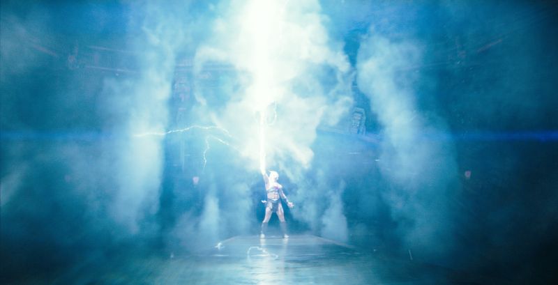Figura recibiendo rayo de energía en escenario lleno de humo.