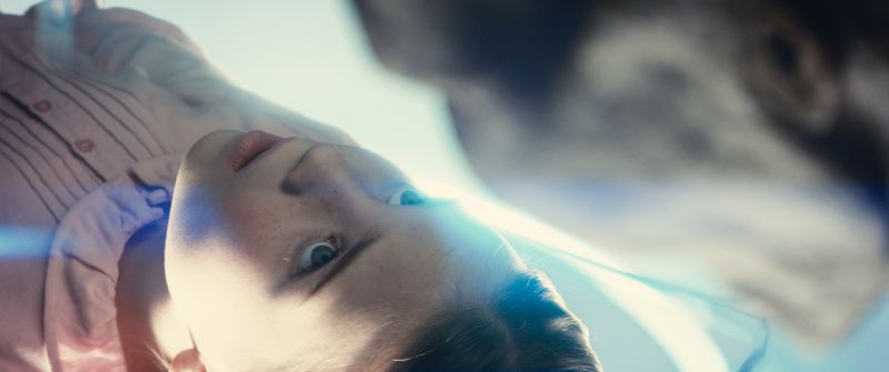 Joven acostado observando de cerca a un extraterrestre en un entorno luminoso.