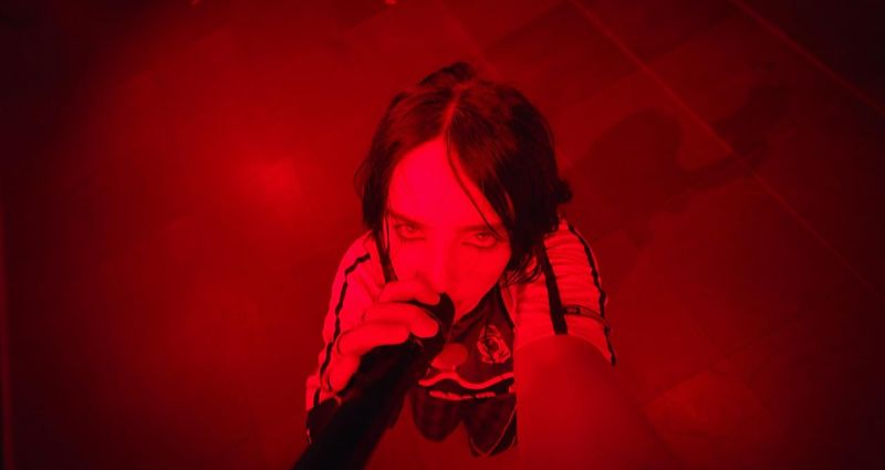 Billie Eilish en escena Hit Me Hard And Soft Primer plano de Billie Eilish cantando bajo una intensa luz roja, mirando directamente a la cámara mientras sostiene el micrófono.