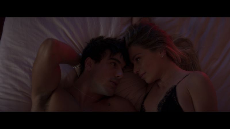 Escena íntima de los personajes de Matías y Lucero acostados, mirándose fijamente bajo una luz tenue y cálida.