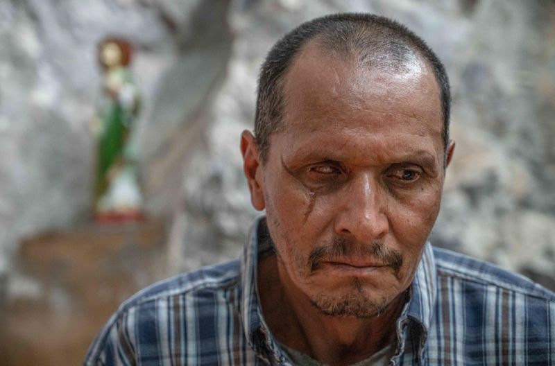 Primer plano del actor Gerardo Trejoluna con una cicatriz en el rostro y expresión severa, con una figura religiosa al fondo.