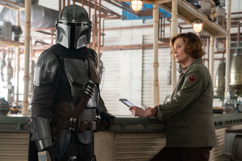 Taller — The Mandalorian and Grogu Din Djarin junto a un personaje en un taller industrial.