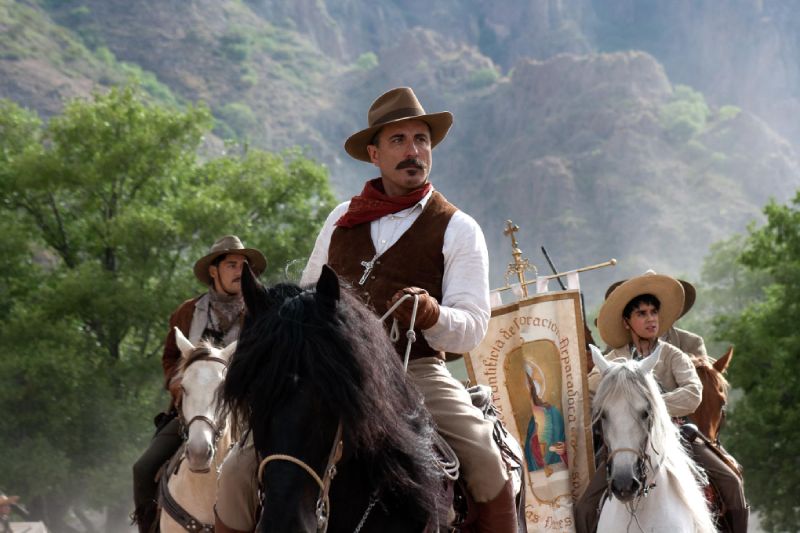 Andy García caracterizado como el General Enrique Gorostieta montando a caballo en el campo junto a tropas cristeras y un estandarte religioso.