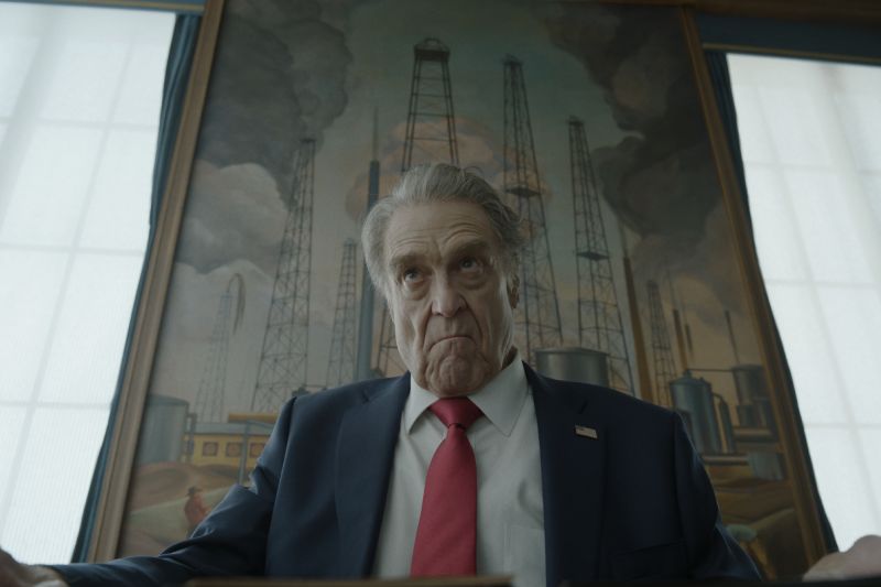 John Goodman como el Presidente Compson mirando fijamente hacia abajo con expresión seria frente a una pintura de torres de extracción petrolera.