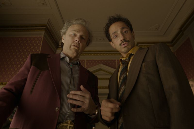 Tom Cruise y Riz Ahmed observando hacia abajo en un ángulo contrapicado en el interior de una elegante habitación decorada.