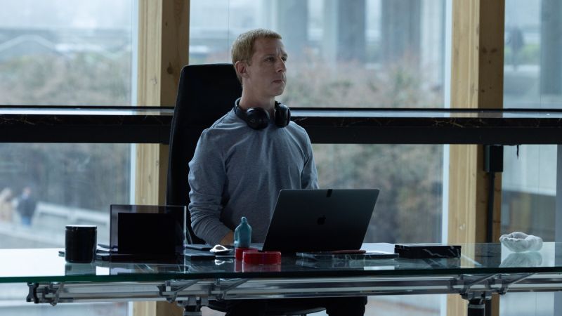 Poder Corporativo en El Poder de la Red Jeremy Strong sentado frente a un escritorio elegante con una laptop y auriculares, manteniendo una expresión seria en un entorno corporativo.