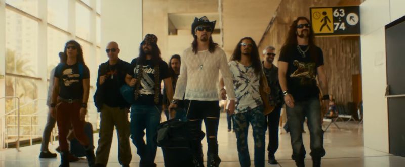 Integrantes de la banda caracterizados para la película Fiesta Pagäna caminando juntos por el pasillo de un aeropuerto con maletas y atuendos de estilo rockero.