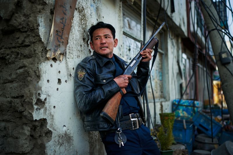 El actor Hwang Jung-min vistiendo chamarra de piel y uniforme cubriéndose contra un muro destruido sosteniendo una escopeta.