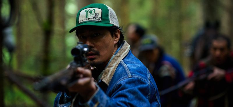 El actor Zo In-sung usando una gorra verde y chamarra de mezclilla apuntando fijamente con un rifle de francotirador en el bosque.