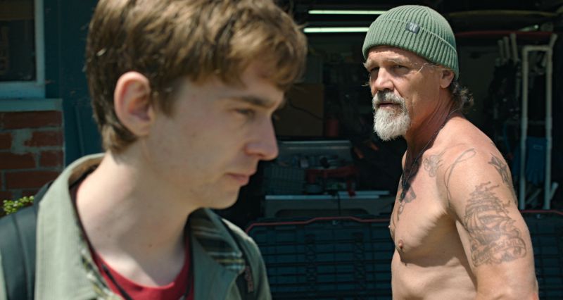 Josh Brolin con gorro tejido y tatuajes mirando seriamente hacia un lado mientras Austin Abrams aparece en primer plano fuera de foco.