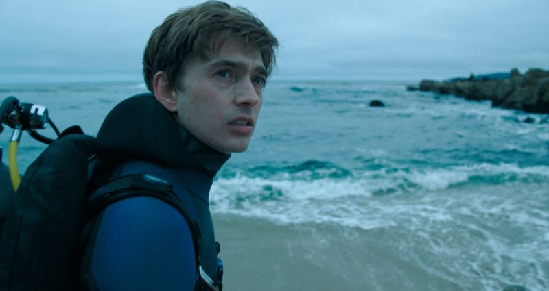 Austin Abrams con traje de neopreno en la orilla de la playa mirando reflexivo hacia el mar bajo un cielo nublado.